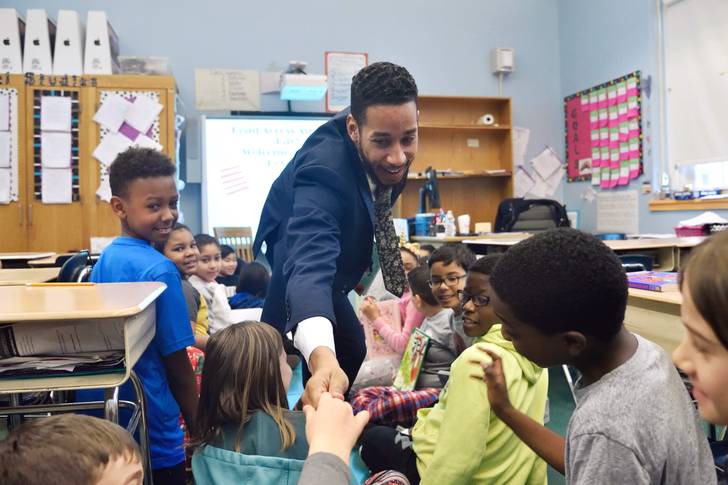 Antonio Reynoso, Councilmember for Brooklyn's 34th Council District, is inside a classroom in March 2018. Reynoso, wearing a suit, crouches to a group of kids, smiling.